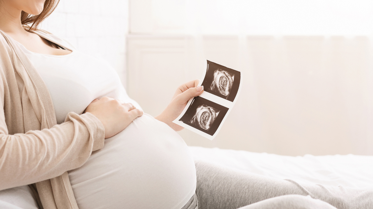 A pregnant woman in white tshirt and beige cardigan holds an ultrasound